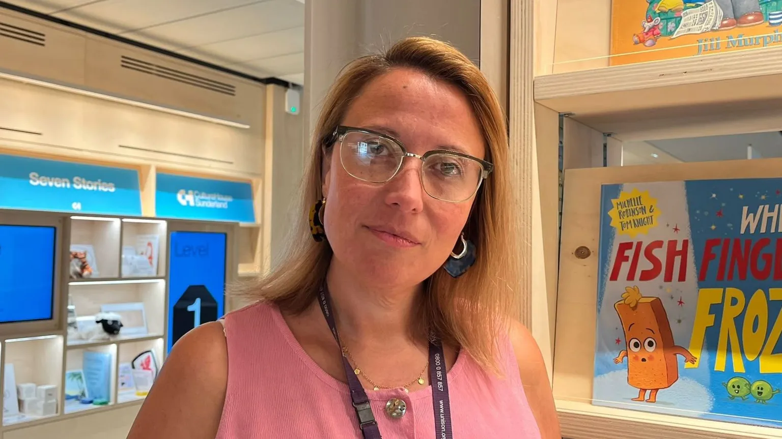 BBC / Kay Davidson Leanne Littlewood is looking at the camera with a neutral expression on her face. She has mid-length blonde hair and is wearing a pink, sleeveless top and glasses. She is standing in front of bookshelves.